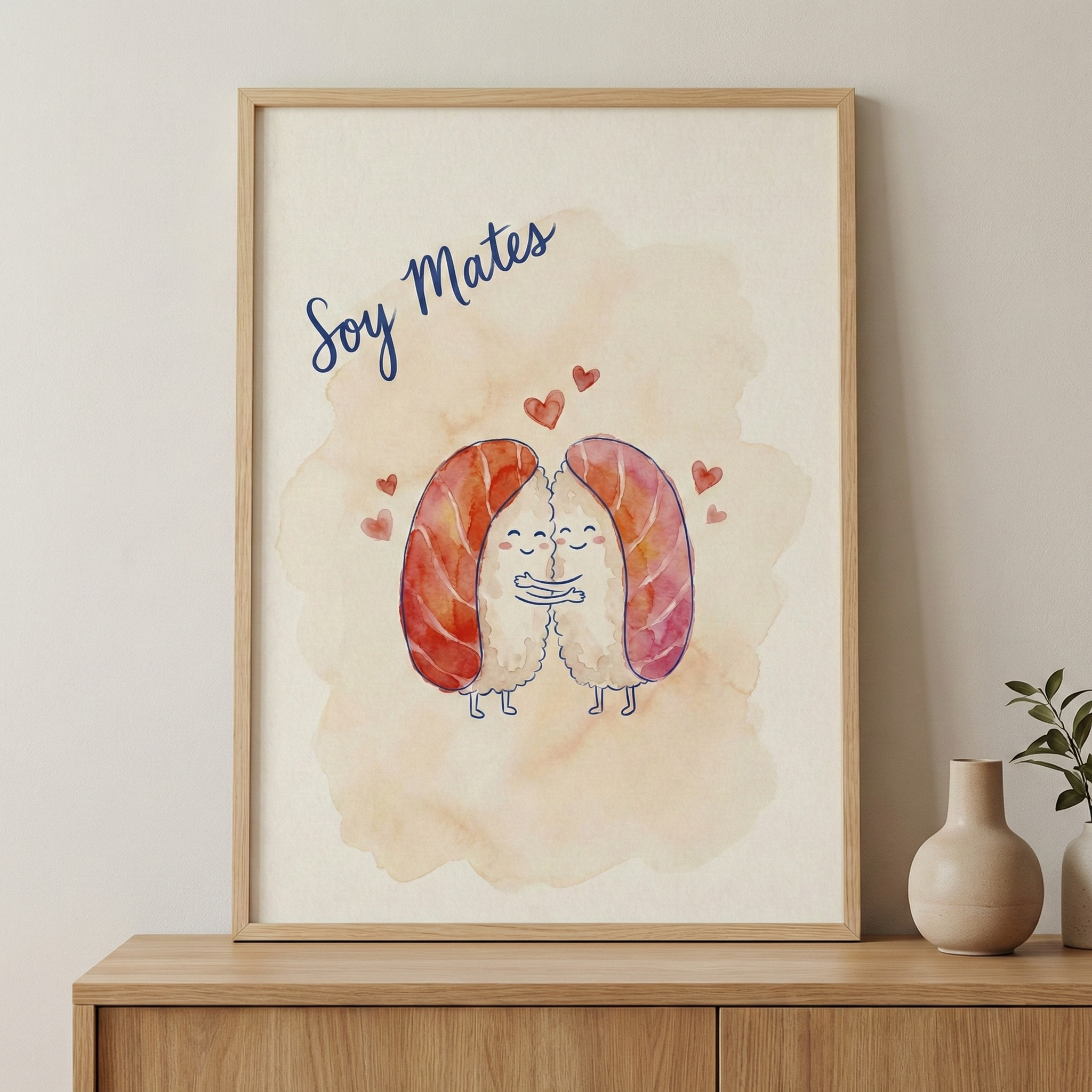 A light wood-framed illustration leans against a neutral-toned wall on a wooden sideboard. The artwork features two smiling, cartoon nigiri sushi characters hugging, surrounded by small red hearts on a soft peach watercolor-style background. The phrase "Soy Mates" is written in a blue cursive font at the top. To the right of the frame are two small ceramic vases, one holding a green leafy branch.