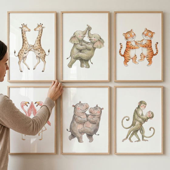 A hand adjusts a sweet dancing animals wall art set of 6 with watercolor giraffes, elephants, tigers, hippos, and monkeys.
