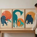 A set of three minimalist art prints in light wood frames sits side-by-side on a wooden surface. Each print features stylized animal silhouettes against a large, circular sun-like backdrop. 
