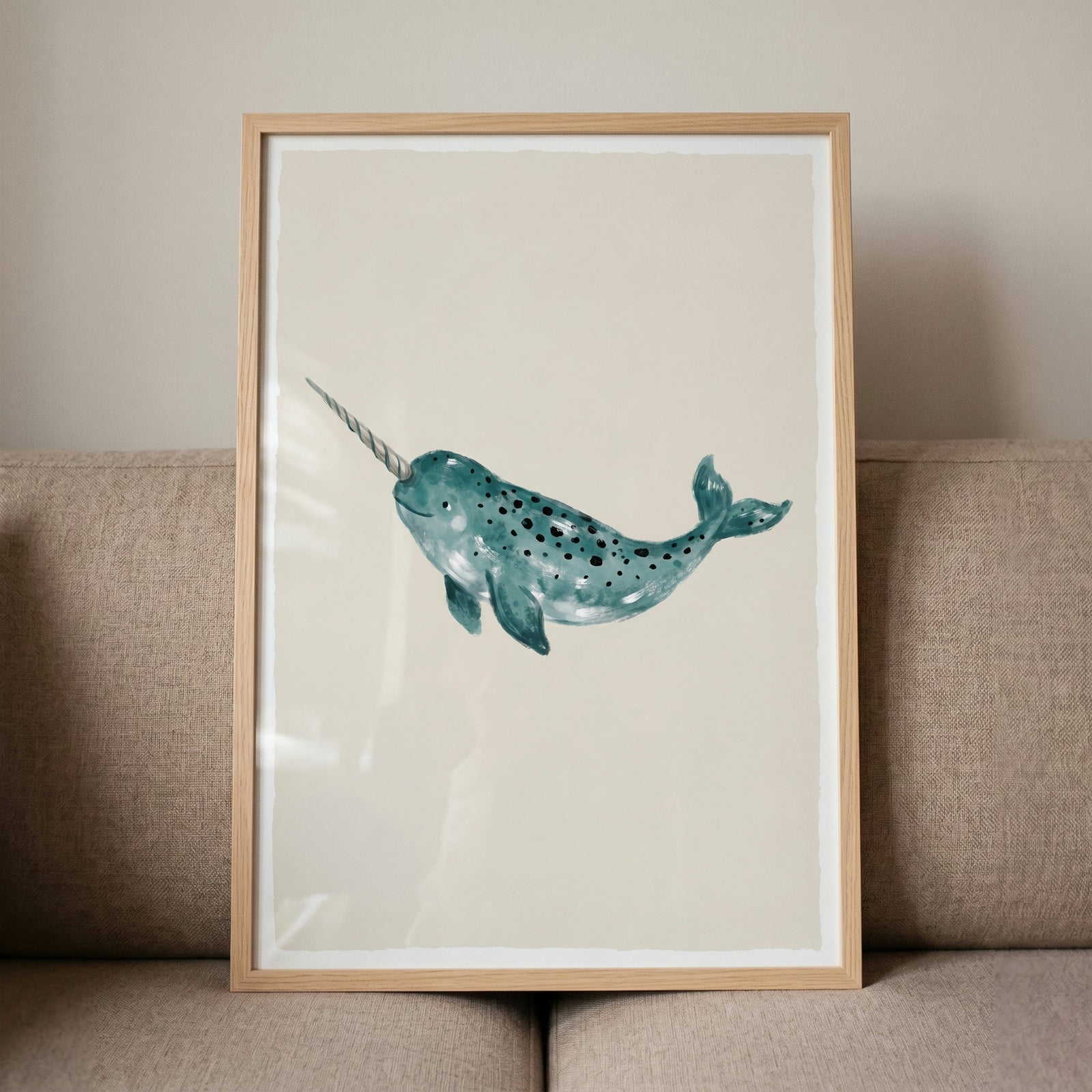 Teal watercolor narwhal wall art with a long tusk and dark spots, framed in light wood against a beige sofa.