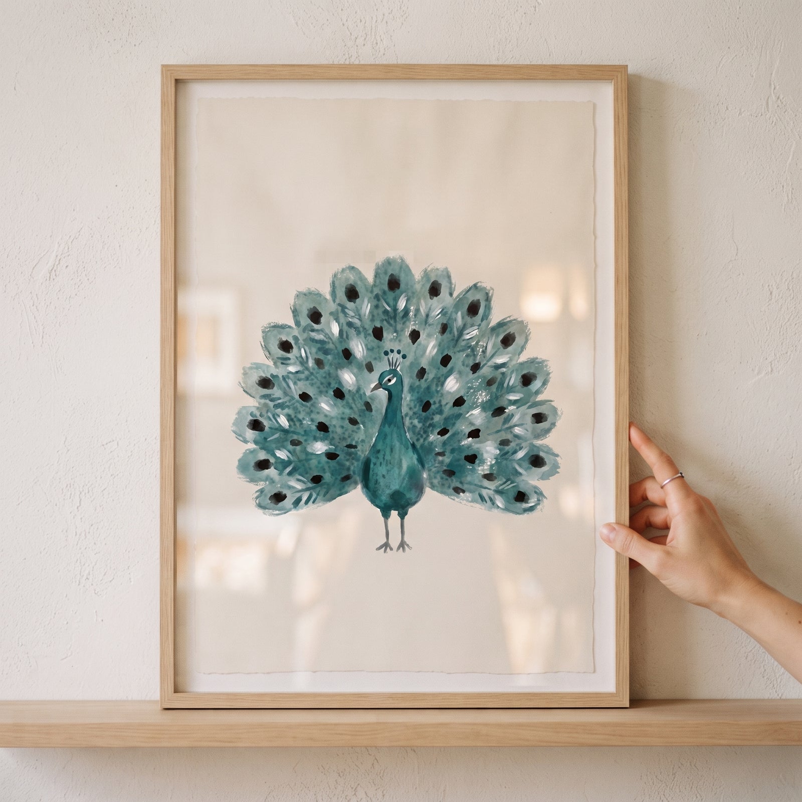 Hand holding a framed teal watercolor peacock wall art with spread tail feathers on a light wood shelf.