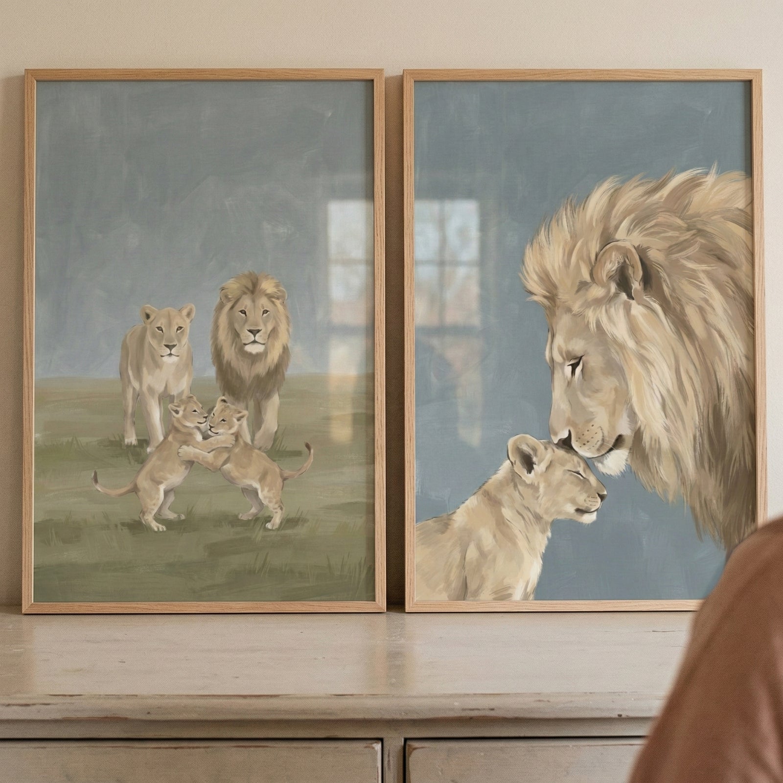 Two framed artworks are displayed side-by-side on a light-colored, rustic wooden surface. Both pieces feature a gentle, illustrative style with a soft color palette of tans, creams, and muted blues.

The artwork on the left depicts a group of lions in a grassy field. In the background, a large male lion with a full mane and a sleek female lion stand watch. In the foreground, two small lion cubs are shown in a playful embrace. The scene is set against a soft green landscape under a hazy, muted blue-grey sky