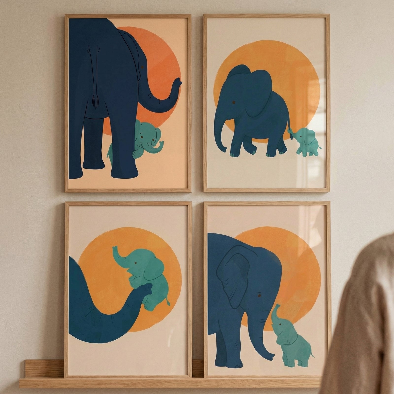 Four framed illustrations are displayed in a grid on a light-colored wall, resting on a wooden shelf. Each illustration features a minimalist, flat-color style with a large blue elephant and a smaller green baby elephant set against a beige background with a large orange circle resembling a sun. 
