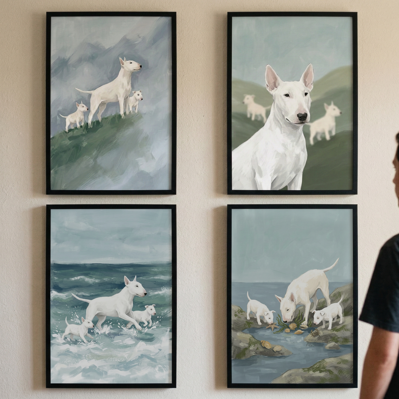 Four black-framed rectangular artworks are arranged in a two-by-two grid on a light beige wall. Each piece features white bull terriers in various natural environments. 

In the top-left illustration, an adult dog and two puppies stand on a grassy hill before a backdrop of hazy mountains. To the right, a portrait-style artwork shows the head and shoulders of a bull terrier with two more dogs in the distance on a soft green slope. 

