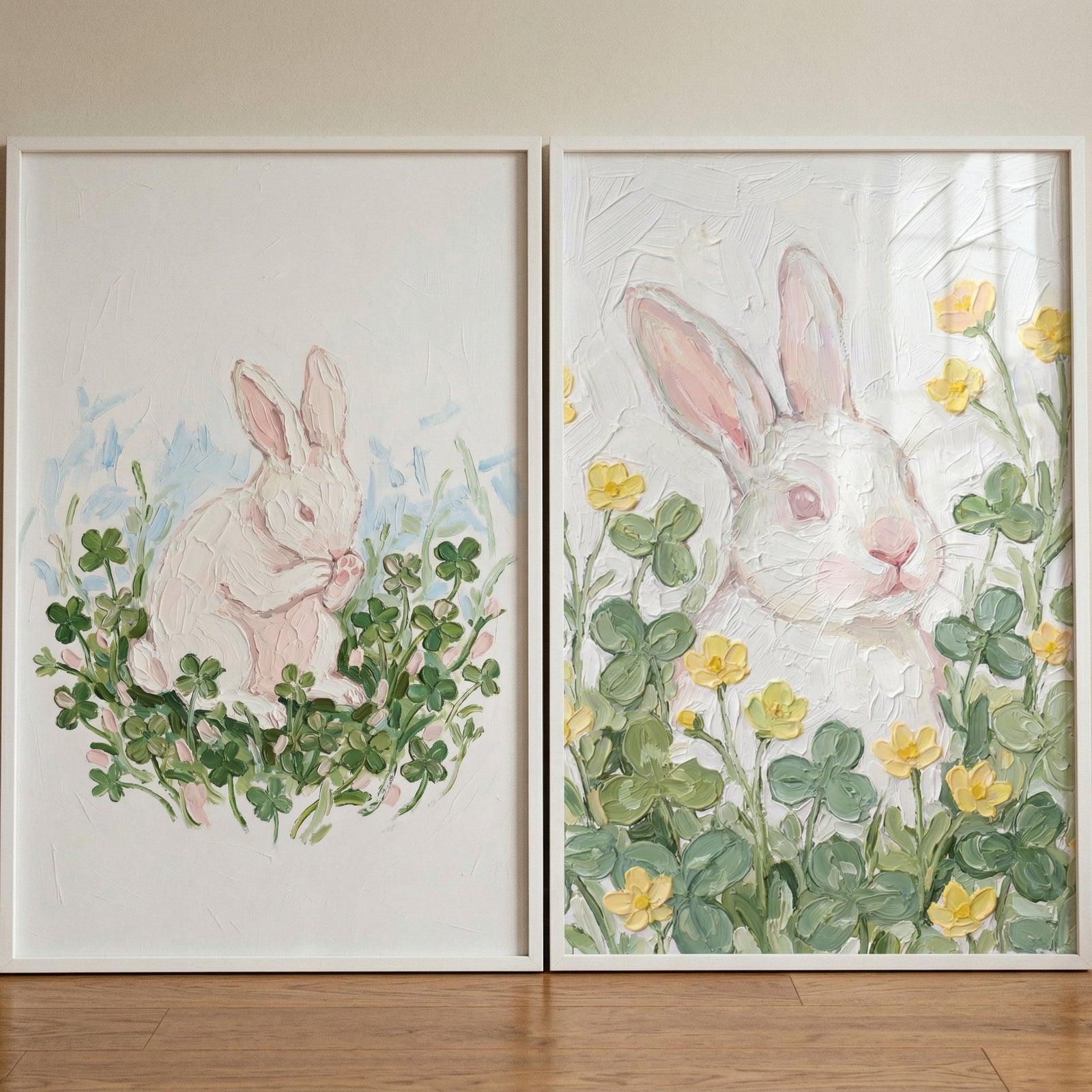 Two framed paintings of a white rabbit are displayed side by side on a wooden floor against a neutral wall. The left painting shows a small rabbit sitting and grooming itself among green clover and soft pink buds, while the right features a close up of a rabbit surrounded by clover leaves and yellow flowers in a textured, pastel style.
