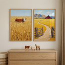 Two framed farm paintings hang above a light wood dresser in a calm neutral room. The left print shows a brown cow peeking out from tall golden grass and the right print shows an open gate and winding dirt road leading to a red barn with mountains behind it.