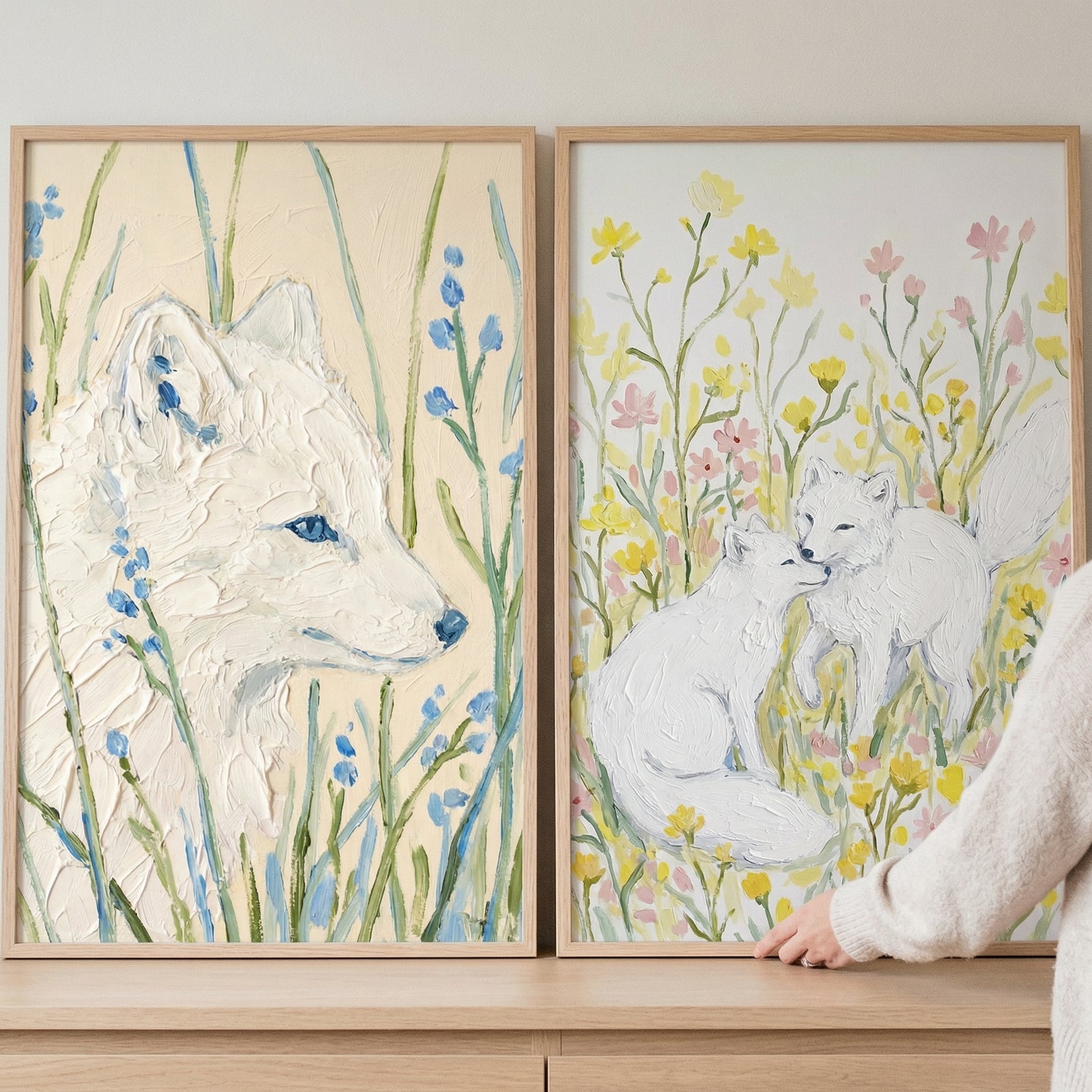 Two textured paintings of white foxes in soft pastel wildflowers, one showing a close up fox with blue accents among tall grass and the other showing two foxes nuzzling in a field of yellow and pink blooms. A hand gently adjusts the framed artwork, suggesting placement in a calm nature inspired room.