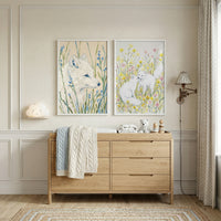 A cozy and bright nursery or child's room features two framed watercolor-style illustrations hanging on an off-white wall with decorative molding. The illustration on the left shows the profile of a single white fox nestled amongst tall blades of grass and delicate blue blossoms. The illustration on the right depicts two white foxes interacting in a field filled with yellow and pink wildflowers.

Below the artwork is a light oak-colored wooden dresser with a minimalist design, serving as a changing station.