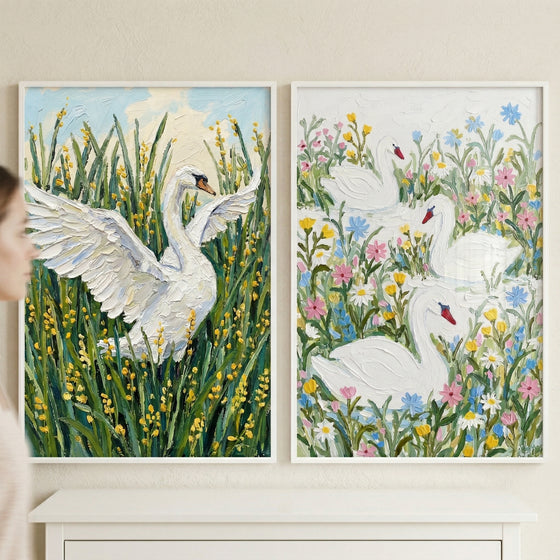 Two framed textured paintings of white swans displayed side by side, one showing a swan with wings spread among tall green reeds and yellow flowers and the other showing three swans swimming among colorful wildflowers. A blurred figure passes by, suggesting the artwork styled in a soft airy home interior.