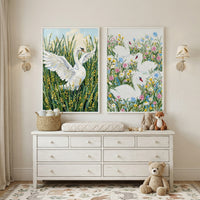 Soft neutral nursery with a white dresser and changing pad beneath two framed swan paintings, one showing a swan with wings open in tall green grass and yellow flowers and the other showing three swans surrounded by colorful blooms. Cozy details like plush toys, a woven basket, wall sconces, and a patterned rug create a gentle welcoming space for a baby.