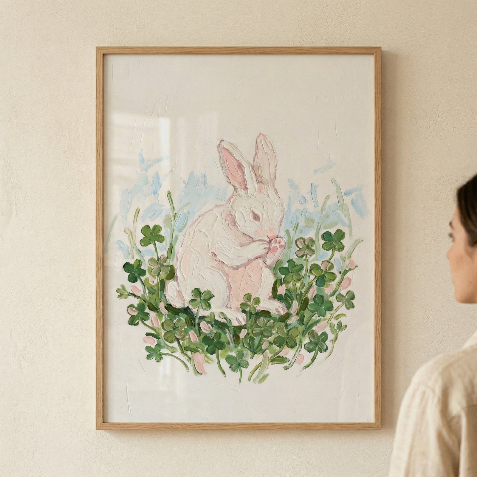 A framed illustration of a white rabbit is shown hanging on a light, textured wall. The animal is depicted sitting in the center of a dense cluster of green clovers, with its paws raised to its face in a grooming gesture. The artwork is rendered with thick, visible strokes that create a textured surface, detailing the rabbit's soft fur and the individual leaves of the plants.
