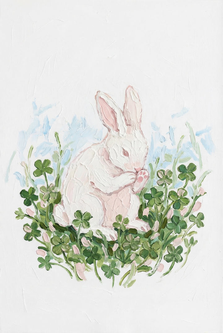 A soft painted white bunny sits curled in a nest of green clover leaves, gently holding its paw near its face. The light background and delicate pink accents give the scene a peaceful, cozy feel.
