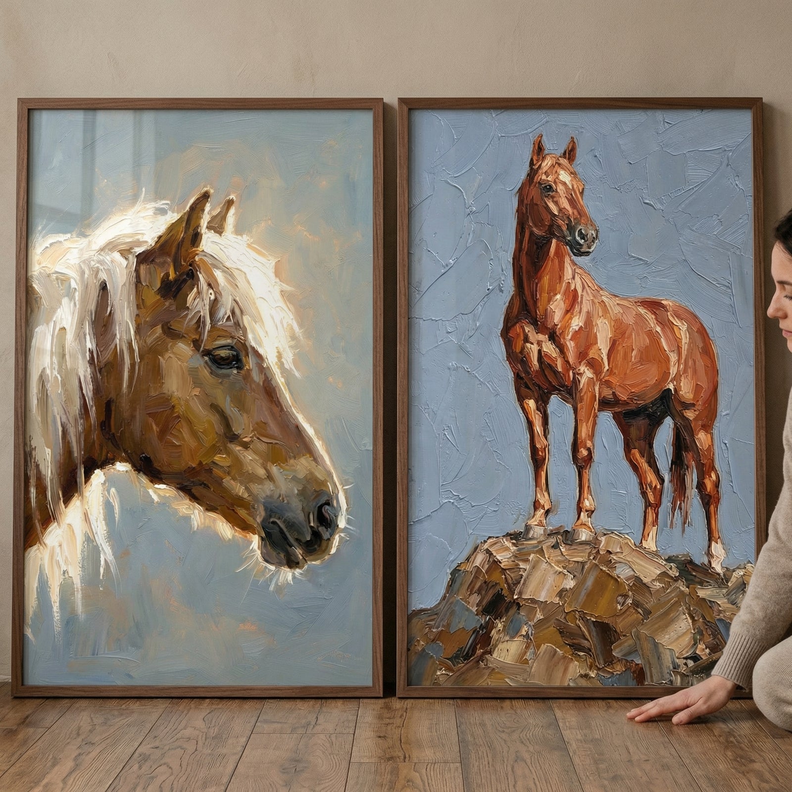 Two framed textured horse paintings displayed on the floor, one showing a close up of a brown horse with a light mane and the other showing a full horse standing on rocky ground against a soft blue background. A person kneels beside the artwork, suggesting placement in a warm rustic interior.