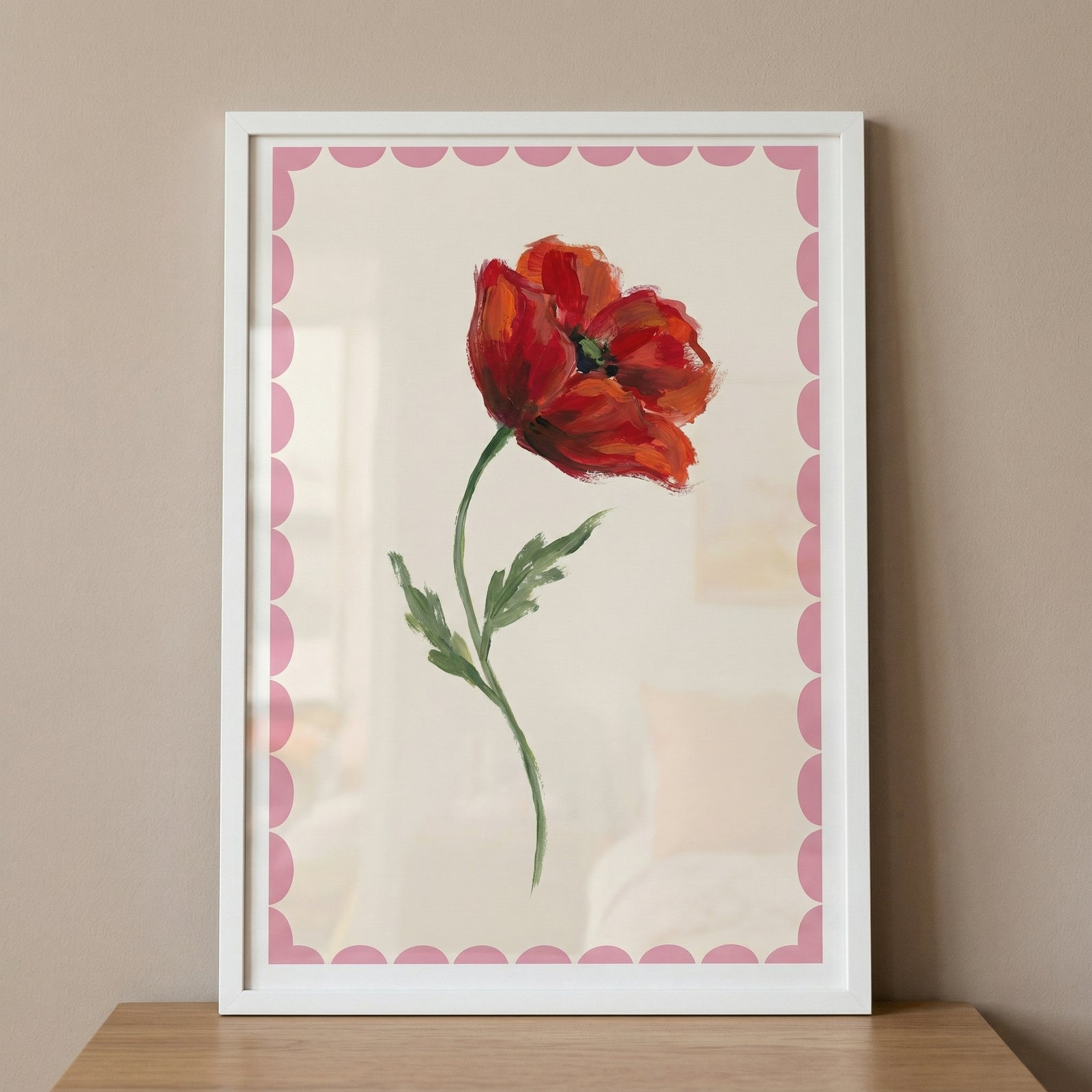 Framed painting of a single red flower with soft green stem and leaves on a light background with a pink scalloped border, resting on a wooden surface. The bold red bloom stands out against the soft tones, creating a simple yet eye catching floral accent.