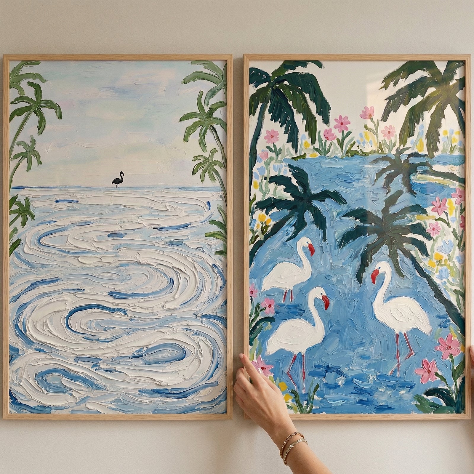 Two framed textured coastal paintings displayed side by side, one showing soft swirling ocean waves with a distant bird and palm trees and the other showing white flamingos standing in blue water surrounded by tropical greenery and pink flowers. A hand adjusts the frame, suggesting styling artwork in a relaxed beach inspired space.