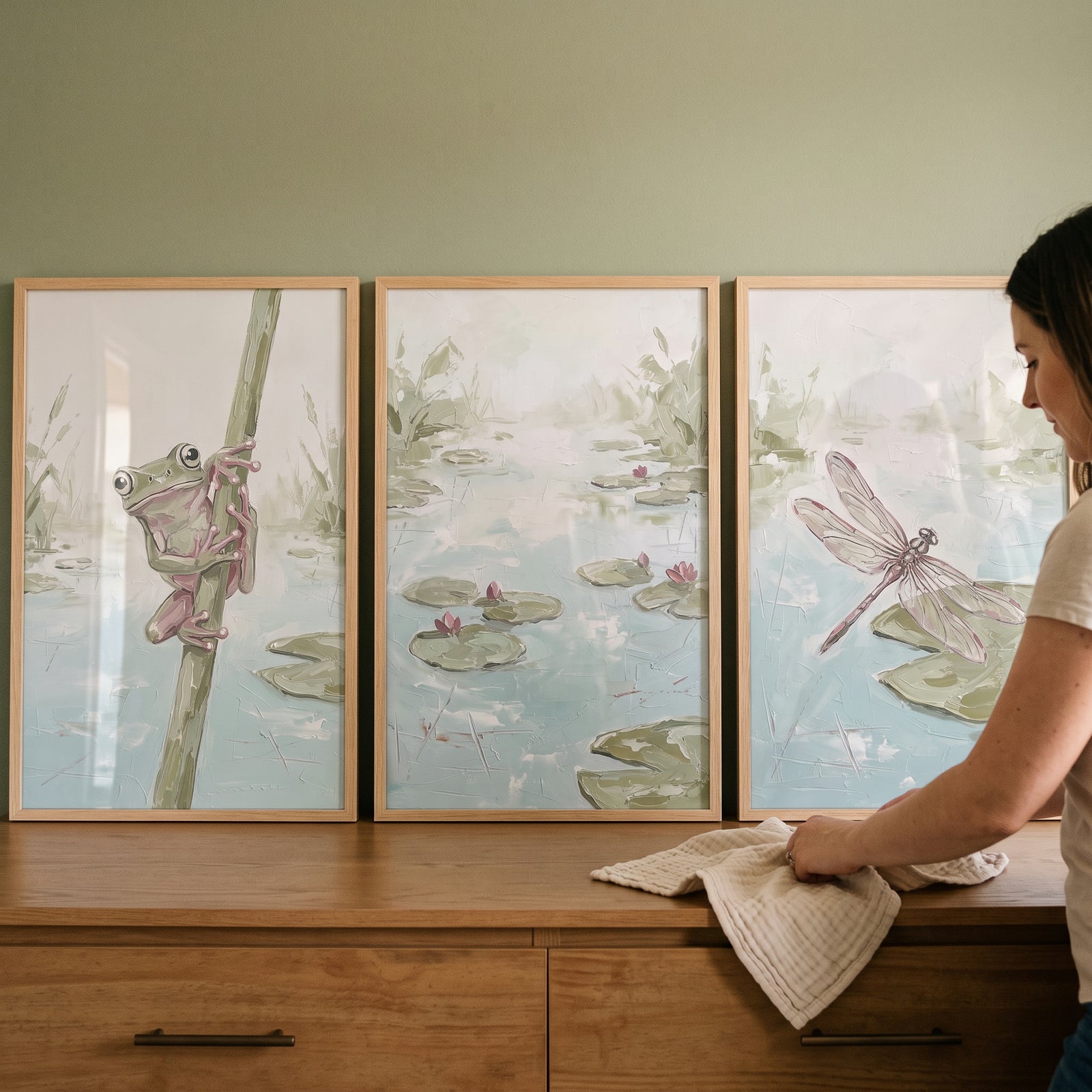 A woman wipes a textured impasto wetland pond wall art set of 3 featuring a frog, lily pads, and a dragonfly.