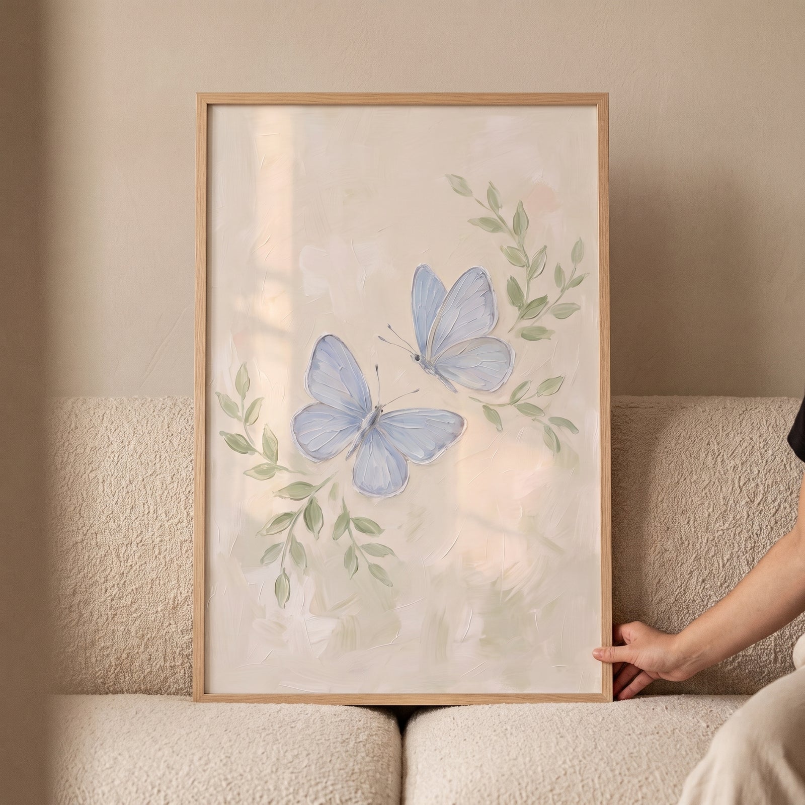 Textured pale blue butterfly artwork with green leaves in a wooden frame, held by a person on a cream sofa.