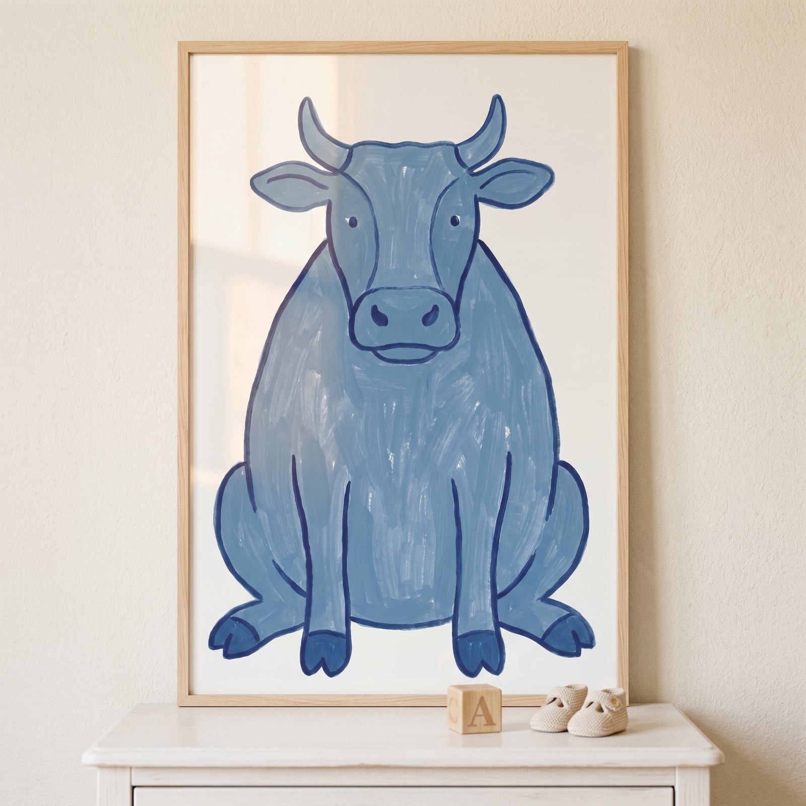 Framed textured slate blue cow wall art sits on a white dresser next to baby shoes and a wooden block.