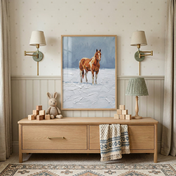 A central framed piece of artwork hangs on a light-colored wall, depicting a brown and white horse standing in a heavily textured, snowy landscape against a soft blue background. The art piece is centered between two wall-mounted sconces with beige shades and brass-colored swing arms.
