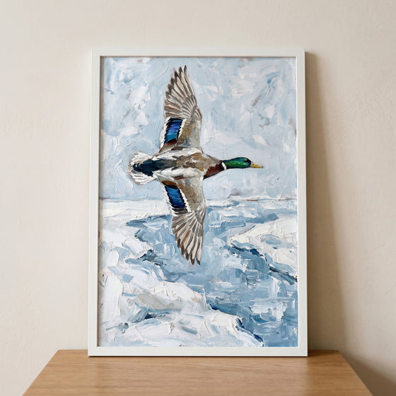 Textured winter mallard duck wall art depicts a bird in flight over icy blue water, displayed in a white frame.