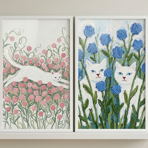 Resting on a white shelf against a beige wall are two white-framed, textured paintings. The artwork features white cats among whimsical yarn flowers: a single cat leaping over a field of pink yarn blossoms on the left, and two cats peeking through a patch of tall blue yarn flowers on the right.