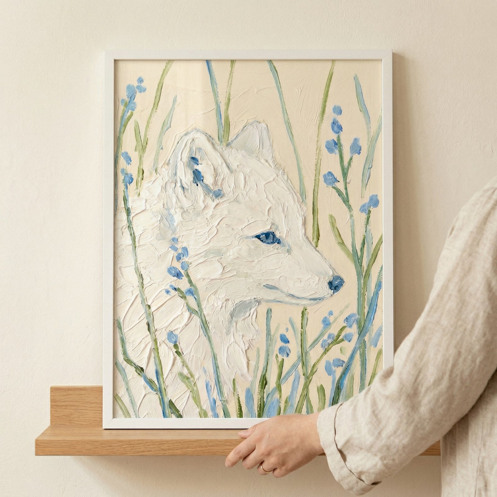 A textured artwork shows the profile of a white fox, crafted with thick, impasto-style strokes that provide a tactile, layered effect. The animal's fur is a mix of creamy whites, set against a neutral beige backdrop. It is surrounded by delicate green stems and small, vibrant blue flowers. The piece is housed in a thin white frame and sits on a light-colored wooden shelf against a plain wall. A person's hand and sleeve are partially visible on the right side.