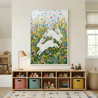 A bright and inviting child's room features a large, vertically-oriented framed piece of textured art as its centerpiece. The artwork depicts two energetic white rabbits leaping through a vibrant meadow of colorful wildflowers in hues of yellow, pink, and blue. 