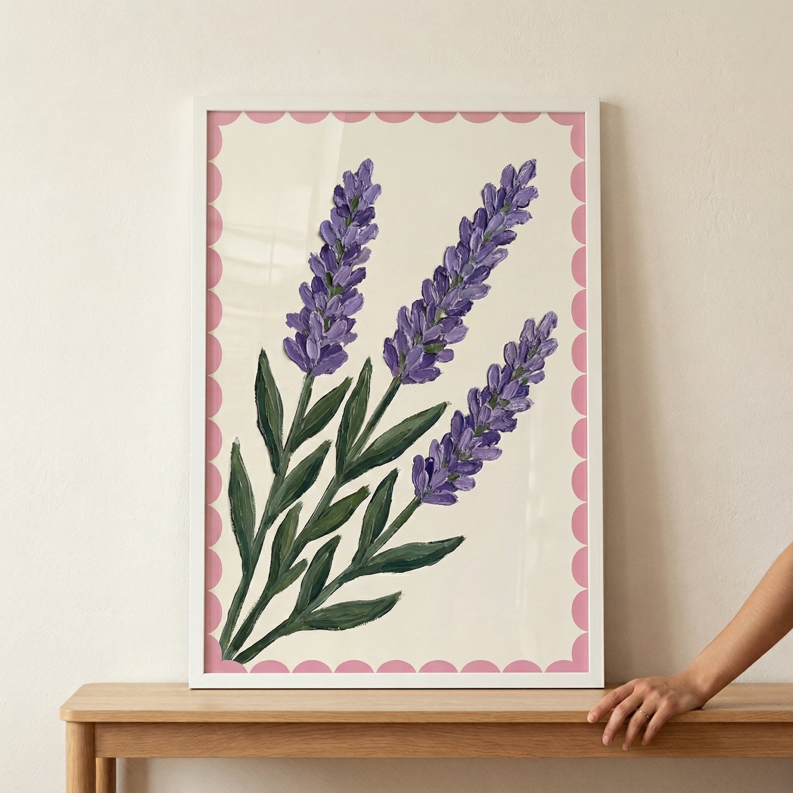 A textured artistic piece features three stems of lavender, created with a technique that shows thick, visible strokes. The lavender blossoms are rendered in shades of purple, while the stems and leaves are various greens. This central image is set against a cream-colored backdrop and framed by a pink scalloped border. The entire work is held within a slender white frame, which rests on a wooden table against a neutral wall. A hand is seen resting near the bottom right corner of the frame.