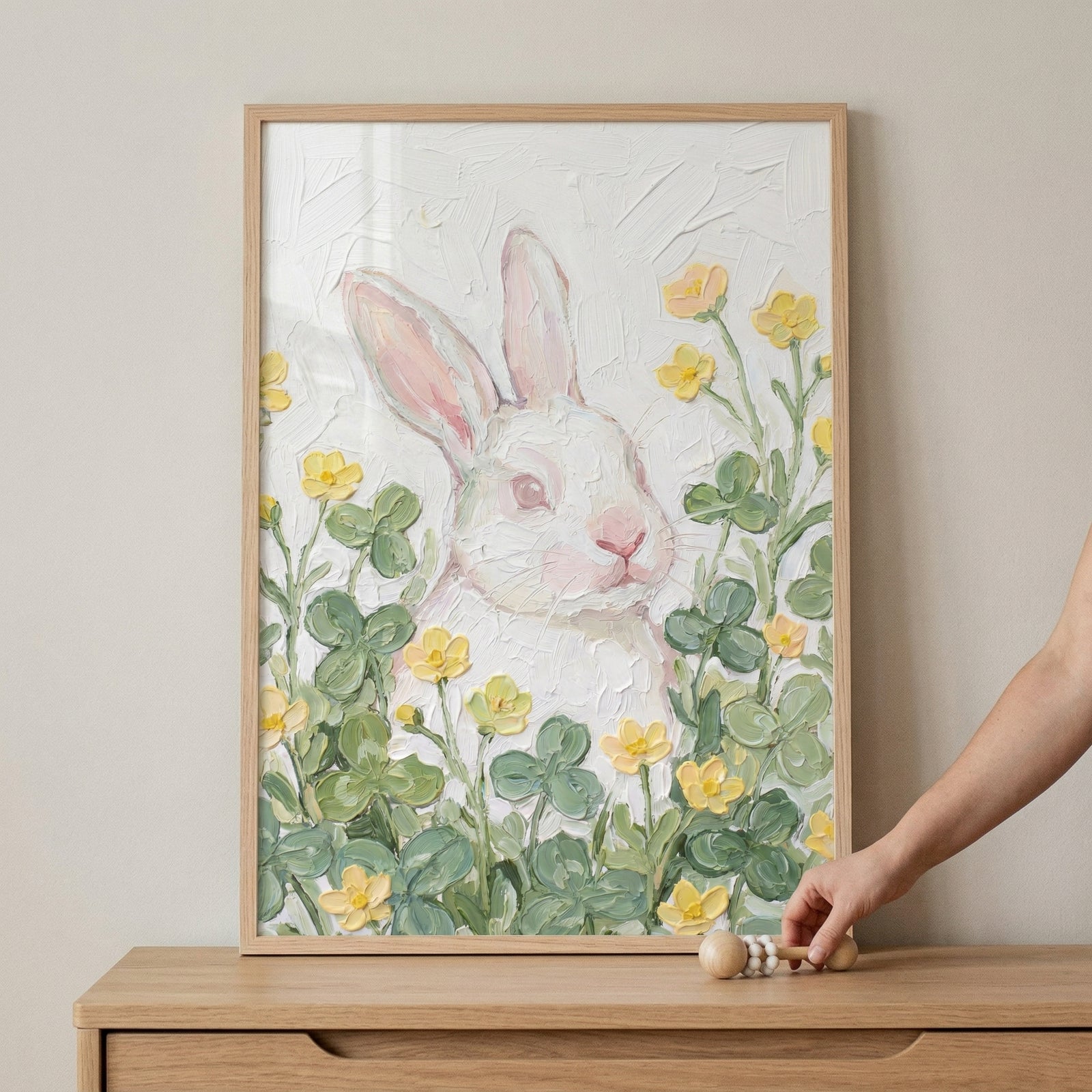 A framed artwork displays a white rabbit with soft pink ears and a pink nose, nestled among green clover-like leaves and small yellow flowers. The illustration features a heavy impasto technique, with thick, textured layers of medium that create a raised, tactile surface for both the animal and the flora. The rabbit is centered against an off-white, textured background. 