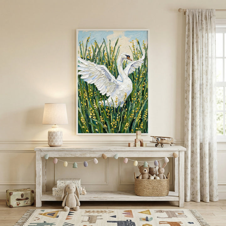 Framed painting of a white swan with wings spread standing in tall green grasses with small yellow flowers hangs above a light wood console in a cozy nursery. Soft decor like a table lamp, garland, woven basket of stuffed animals, and neutral textiles creates a calm and inviting space around the artwork.