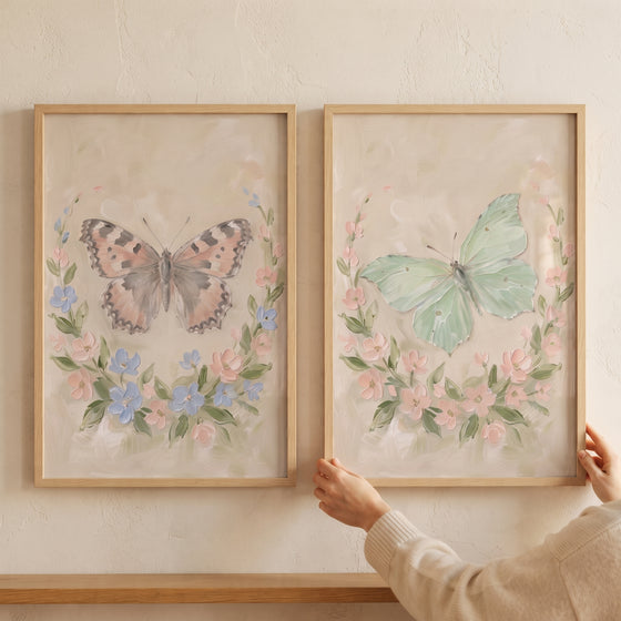 Hands hold a thick oil floral butterfly wall art set of 2 in wood frames with pink and green butterflies and flower wreaths.