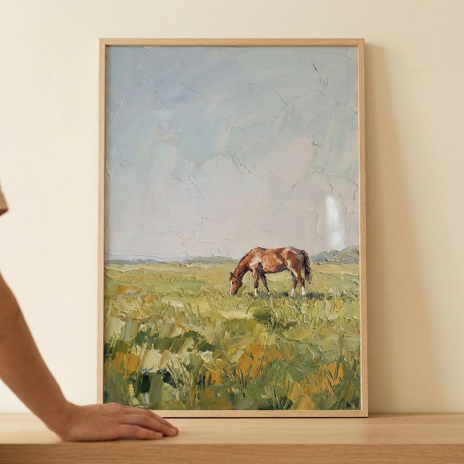 A vertical framed artwork showcases a solitary brown horse grazing in a vast, open field. The horse, with its reddish-brown coat, is the central figure, standing amidst tall, textured grass rendered in a palette of green, yellow, and earthy ochre.

