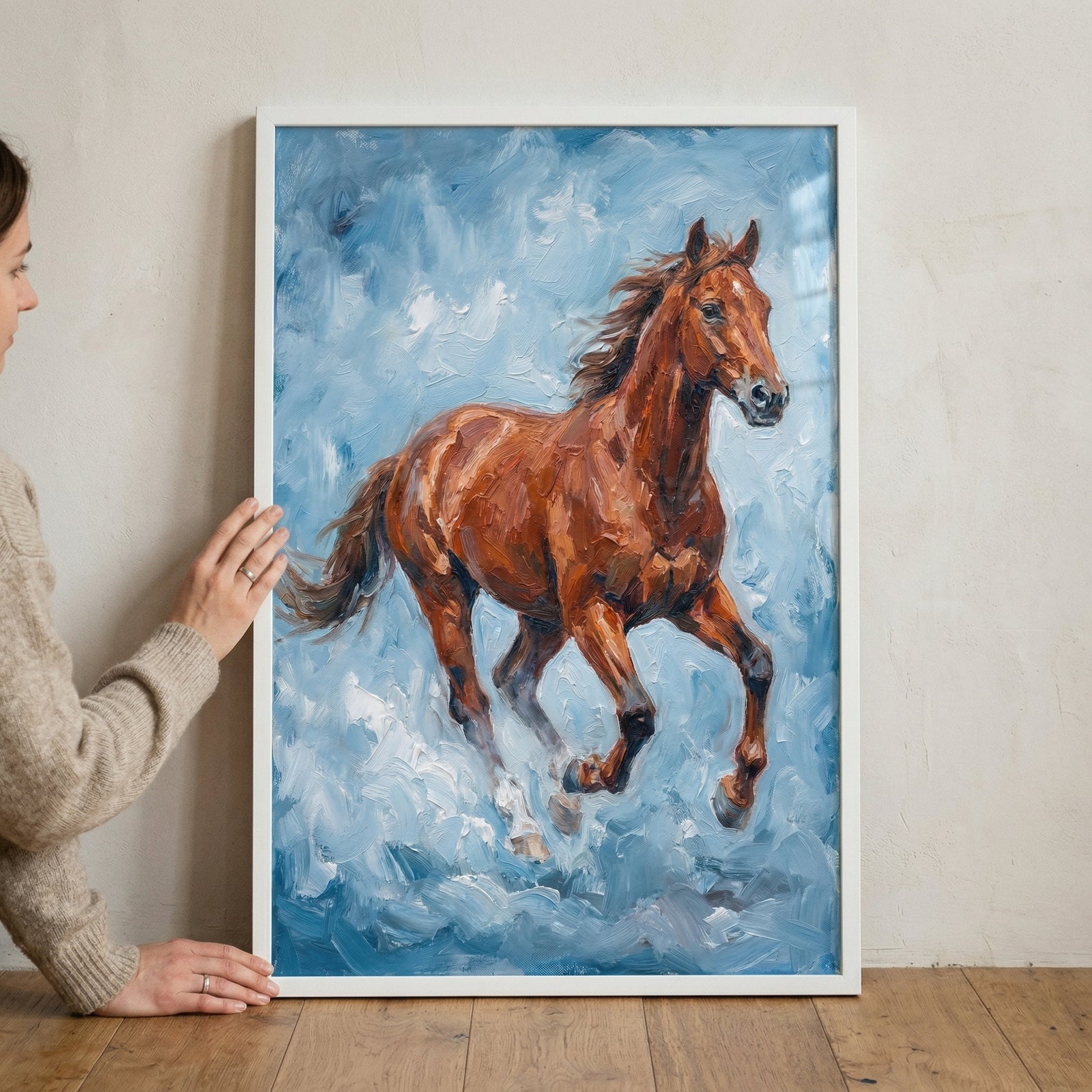 A person wearing a beige knit sweater holds a white-framed artwork featuring a galloping brown horse. The artwork is characterized by thick, expressive brushstrokes that create a textured, impasto effect. 