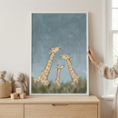 An illustration of three giraffes—two adults and one smaller calf—is featured in a vertical white frame resting on a light wood dresser. The artwork uses soft colors, with the giraffes shown against a textured blue background and green brushstrokes representing grass at the bottom. To the left of the frame, a plush rabbit and a small plush bear in a woven basket sit next to three wooden blocks. A hand is visible touching the side of the frame. The setting is a bright room with neutral walls.