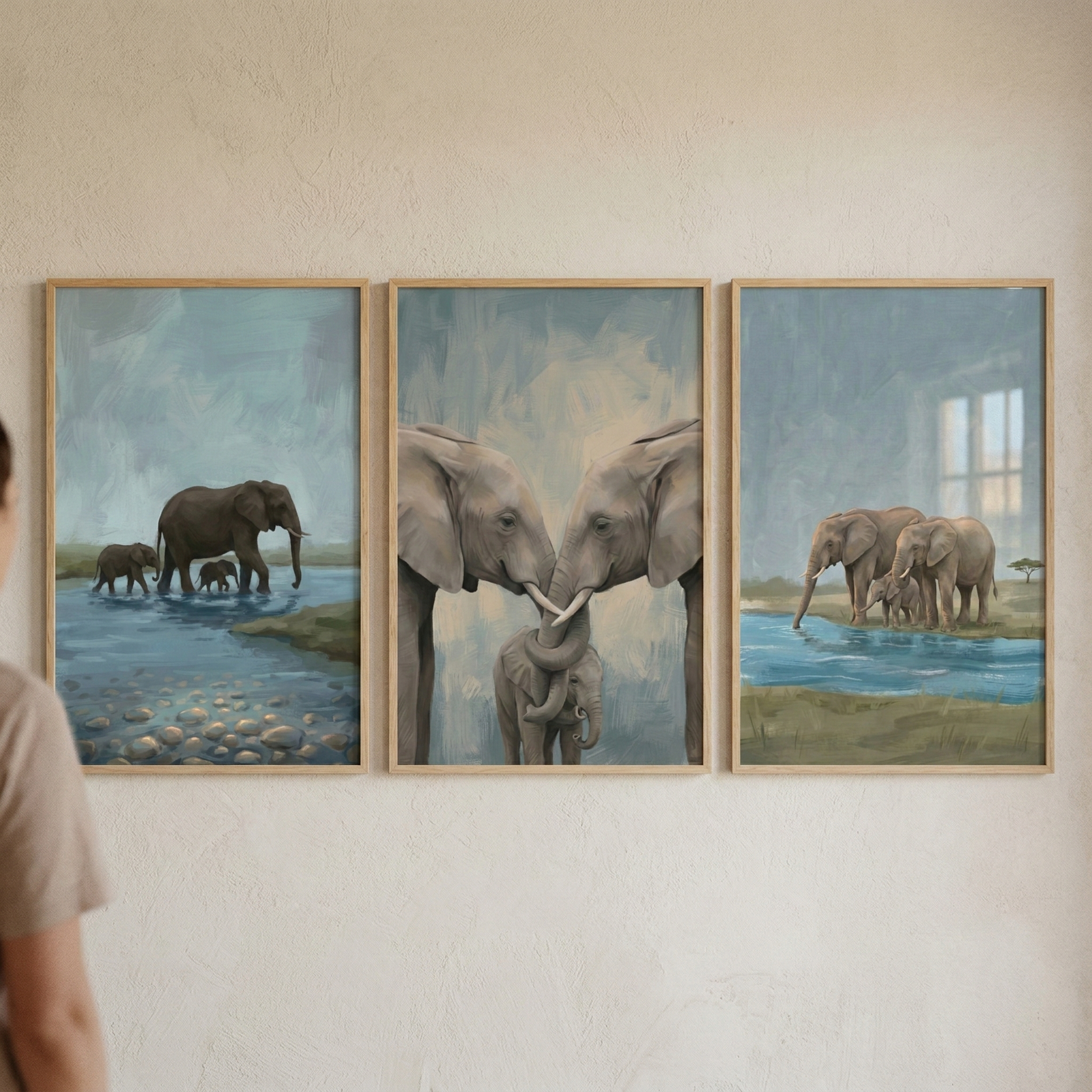 Three framed elephant paintings hang side by side on a light wall in soft muted colors. One artwork shows a mother elephant walking through shallow water with two calves while smooth stones are visible beneath the surface. Another shows two adult elephants gently touching trunks above a baby elephant, and the third shows a small group of elephants gathered beside a calm blue watering hole.