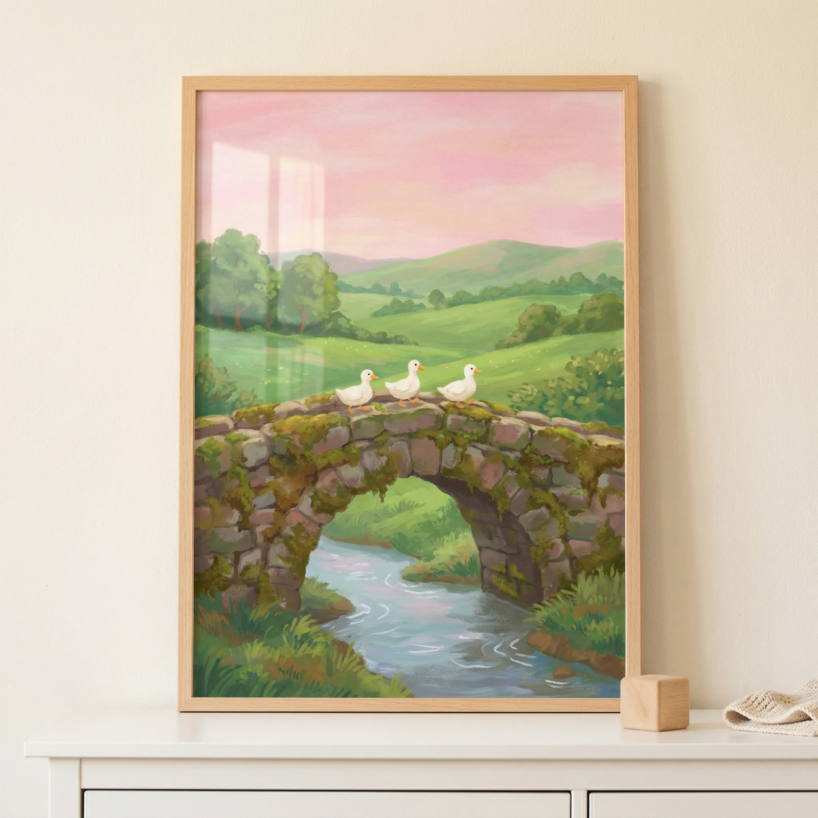 This vertical framed illustration depicts a tranquil pastoral scene. In the center, three small white ducks with orange beaks are walking in a line across an old, moss-covered stone arch bridge. Below the bridge, a clear stream winds through grassy banks. The background features soft, rolling green hills dotted with trees under a hazy, sunset-hued sky of pink and orange. The artwork is characterized by its soft textures and a gentle, whimsical feel. 