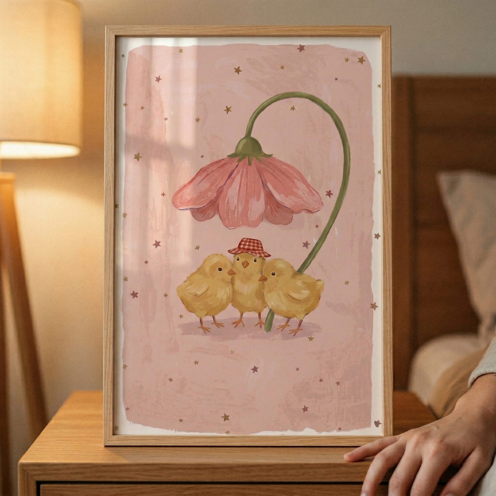 A framed illustration features three small, fluffy yellow chicks standing close together. The chick in the middle wears a tiny red and white checkered hat. Above them, a single large pink flower on a long, curved green stem hangs like an umbrella. The background of the artwork is a muted pink with a soft, textured appearance, adorned with small dark brown stars. The piece is held in a simple wooden frame and rests on a wooden table next to a glowing bedside lamp. 