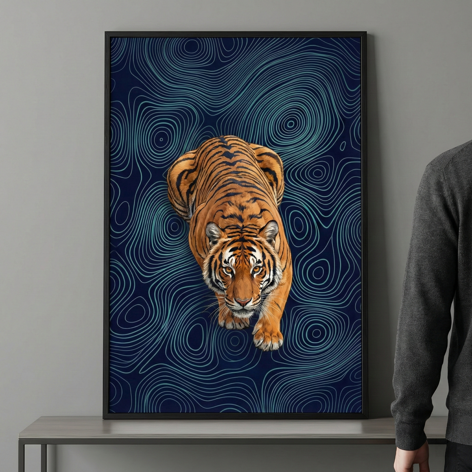 A vertically oriented, framed art piece hangs on a neutral grey wall above a simple table. The artwork features a detailed tiger viewed from an elevated perspective, looking directly forward. Its vibrant orange fur and black stripes stand out against a deep navy blue background filled with fine, light blue concentric and winding lines that create a topographical or rippling effect. A thin black frame surrounds the image. 