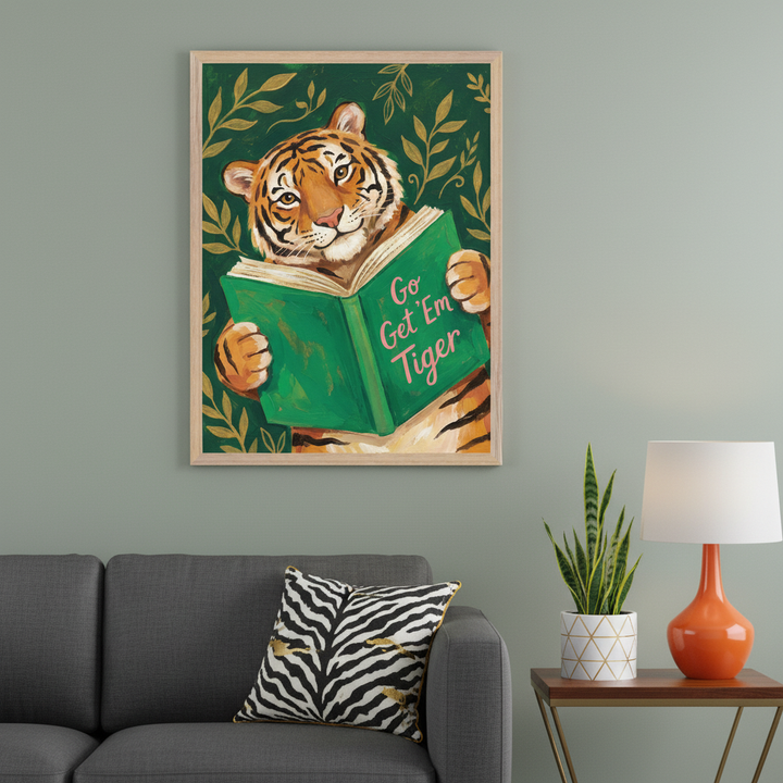 Modern living room with a framed painting of a tiger reading a green book that says "Go Get 'Em Tiger" hanging above a gray sofa. A zebra print cushion rests on the sofa, and a side table with an orange lamp and snake plant is to the right.