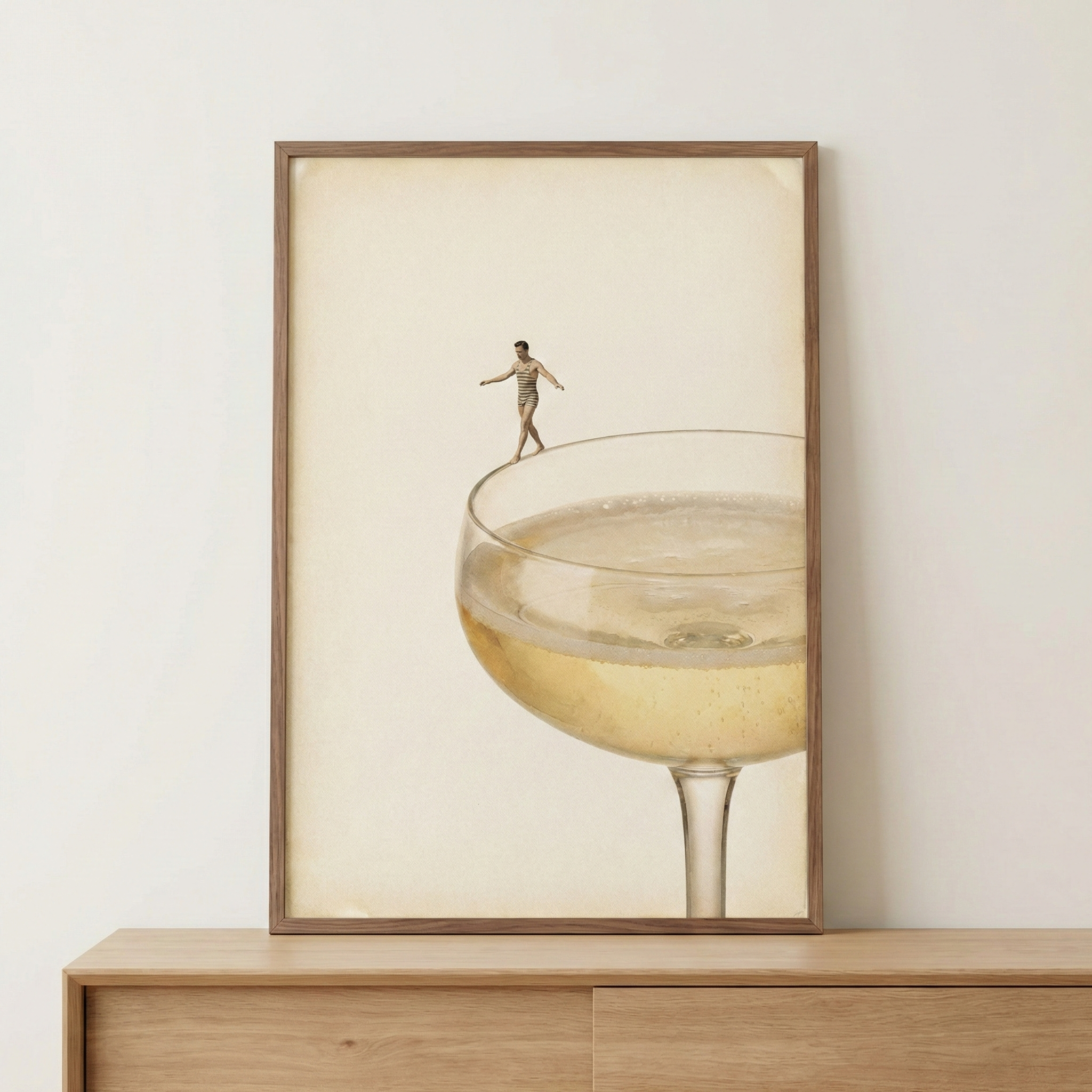 A framed artwork rests on a light wooden surface against a neutral wall. The minimalist composition features a large, elegant glass filled with a bubbly, golden liquid. A miniature figure of a man in vintage-style swimwear is shown balancing on the very edge of the glass's thin rim. The piece has a surreal, vintage aesthetic with a muted color palette of cream, tan, and soft gold tones against a solid, parchment-like background.