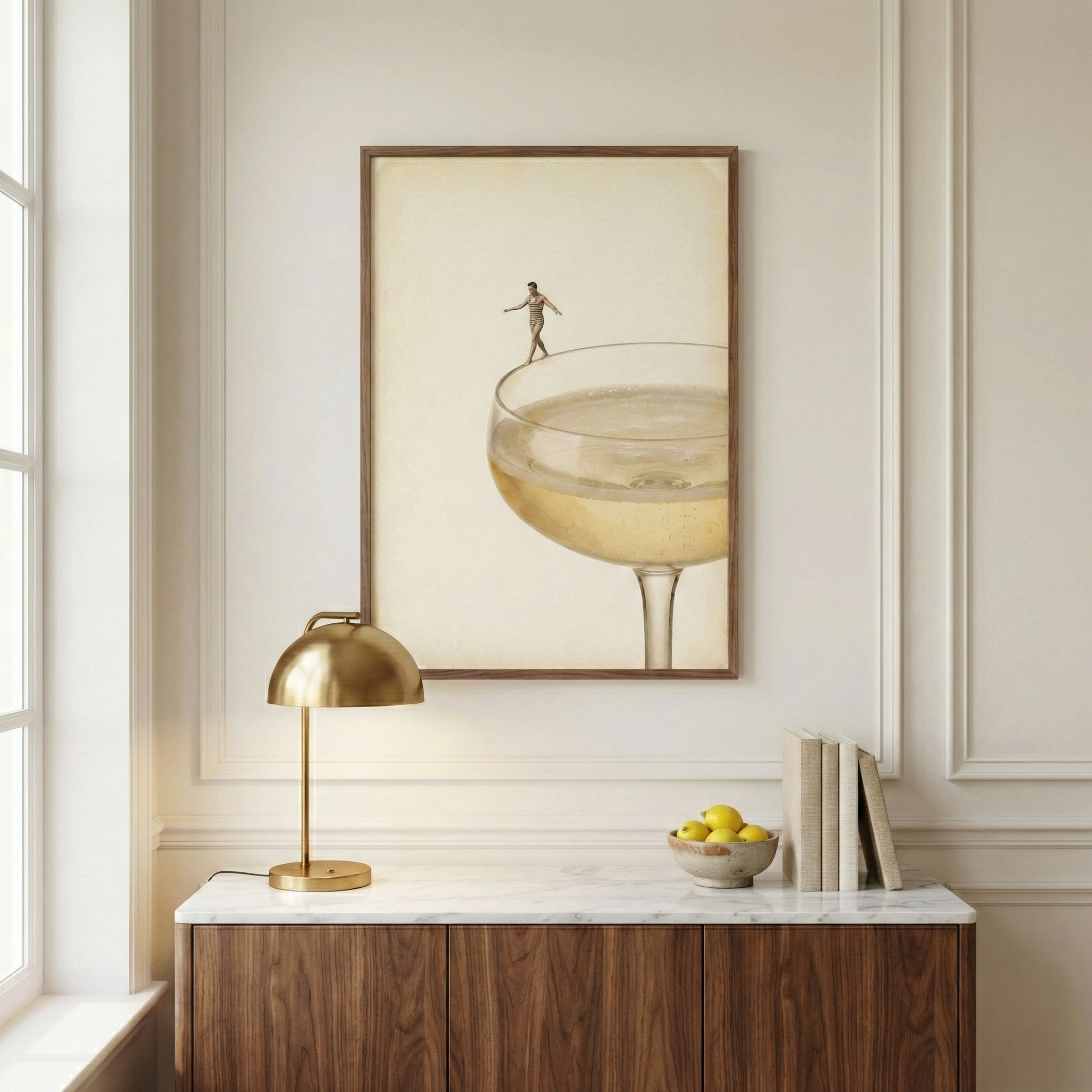 A sophisticated interior features a framed artwork on a neutral wall with elegant molding. The artwork depicts a tiny person walking on the rim of a large coupe glass filled with a sparkling yellow beverage against a cream background. Below, a dark wood console with a white marble top holds a brass dome lamp, a bowl of yellow lemons, and several books. Soft light from a window on the left brightens the scene, creating a calm and modern aesthetic.