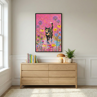 An interior shot of a bright room featuring a large, vertically-oriented framed artwork centered on an off-white wall. The artwork depicts a tortoiseshell cat with mottled black, orange, and cream fur walking toward the viewer through a vibrant garden of purple, yellow, and blue wildflowers. The scene is set against a saturated pink background with colorful speckles, and the cat’s paws are stepping into a shimmering, multi-colored reflection at the base of the frame.
