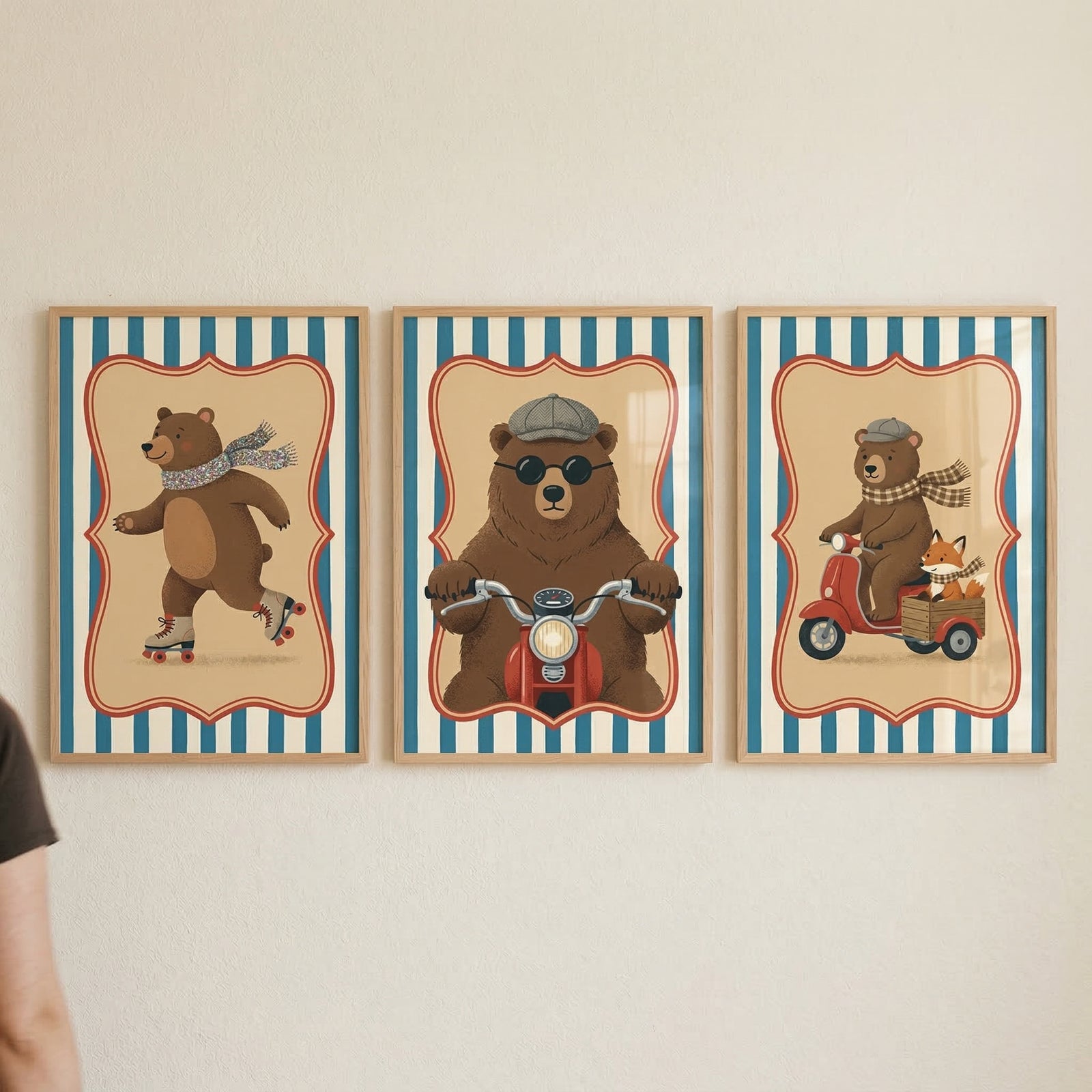 Three framed illustrations are mounted side-by-side on a plain wall. Each artwork features a central scene within a red and gold scalloped frame, surrounded by a blue and white striped border. The left illustration shows a brown bear wearing a gray scarf and roller skates. The center image depicts a bear in a cap and sunglasses riding a red motorcycle towards the viewer. The right frame features a bear on a red scooter with a small fox in a wooden sidecar, both wearing matching checkered scarves.