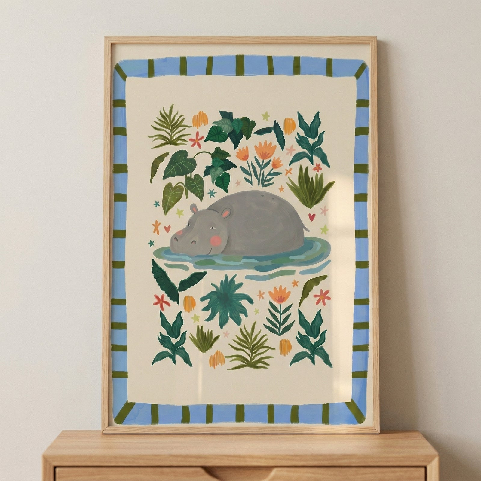 An illustration of a grey hippo in a simple, whimsical style, framed in light wood. The hippo is shown floating in a light blue pool, centered on a cream-colored background. Surrounding the animal are various stylized green tropical plants, palm fronds, and small orange flowers. The piece features a decorative border of light blue with thin vertical green stripes. The framed artwork sits on a wooden surface against a neutral, light-colored wall.