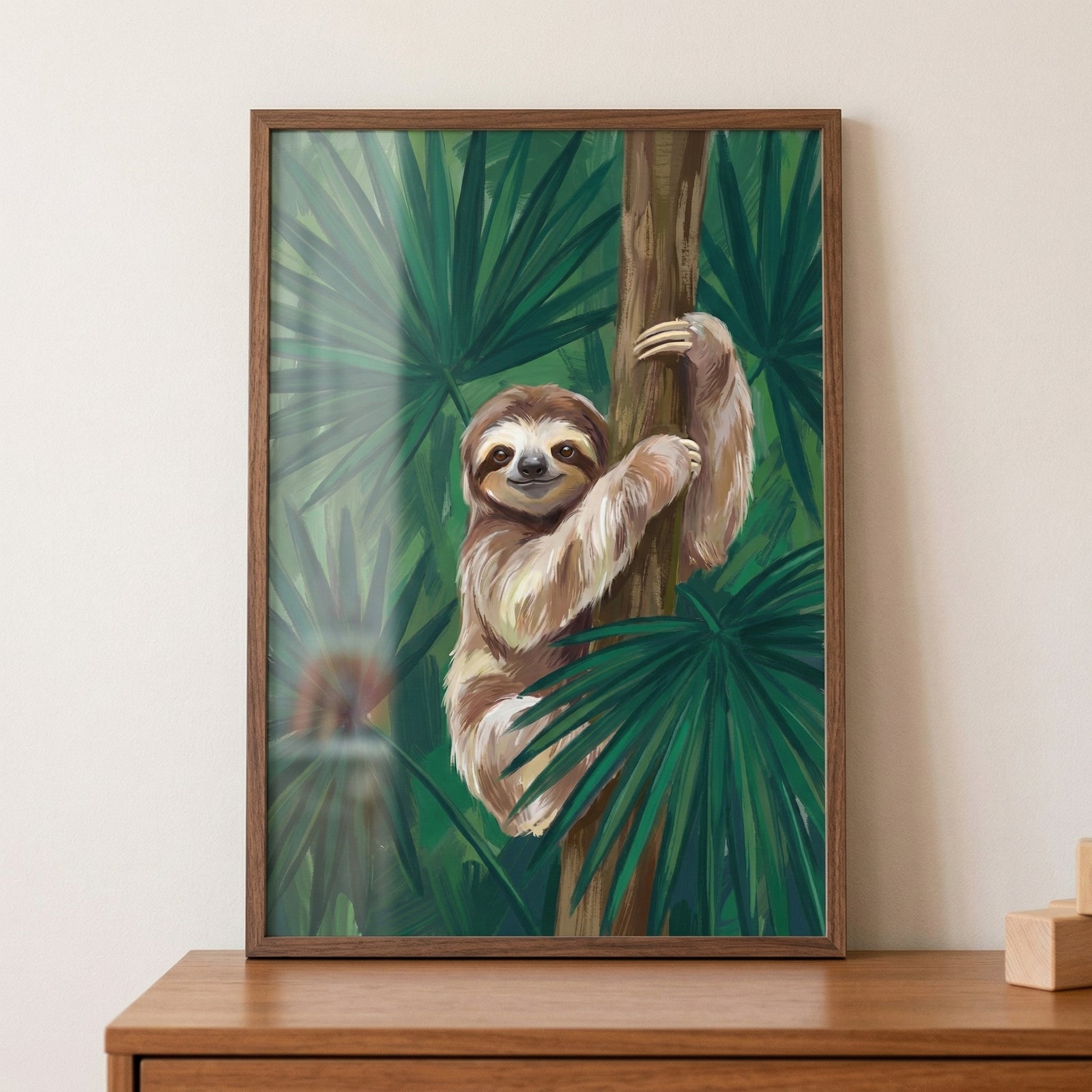 Framed painting of a smiling sloth hugging a tree trunk surrounded by lush green tropical leaves, resting on a wooden surface against a neutral wall. The artwork creates a calm and playful jungle inspired accent with soft light filtering through the foliage.