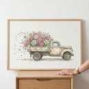 A framed watercolor painting shows a vintage beige truck overflowing with pink and white flowers, with petals gently blowing out the back. A person’s hand rests on the edge of a wooden dresser beneath the artwork.