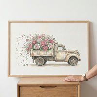 A framed watercolor painting shows a vintage beige truck overflowing with pink and white flowers, with petals gently blowing out the back. A person’s hand rests on the edge of a wooden dresser beneath the artwork.