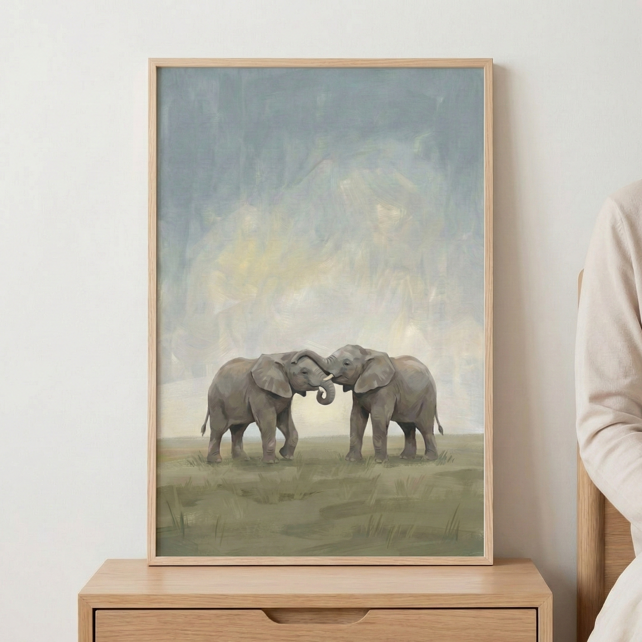 A vertical framed piece of art featuring two elephants standing in a grassy field. The elephants are centered, facing one another with their trunks intertwined in a gentle gesture. The background consists of an atmospheric, hazy sky with soft, blended strokes of blue, gray, and muted yellow. The ground is rendered in earthy tones of olive green and brown. The artwork is held in a light-toned wooden frame and is displayed on a wooden furniture piece against a plain wall. 