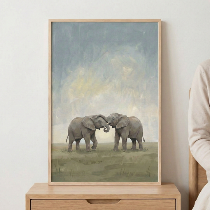 A vertical framed piece of art featuring two elephants standing in a grassy field. The elephants are centered, facing one another with their trunks intertwined in a gentle gesture. The background consists of an atmospheric, hazy sky with soft, blended strokes of blue, gray, and muted yellow. The ground is rendered in earthy tones of olive green and brown. The artwork is held in a light-toned wooden frame and is displayed on a wooden furniture piece against a plain wall. 