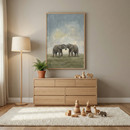 A neutral nursery with a large framed art piece showing two elephants touching trunks in a field. Below, a light-colored wood dresser holds a small plant and wooden toys. A tall floor lamp stands to the left. On the floor, a textured rug is scattered with more wooden playthings, including animal shapes and stacking rings. The room is decorated in soft, warm tones of beige and natural wood.
